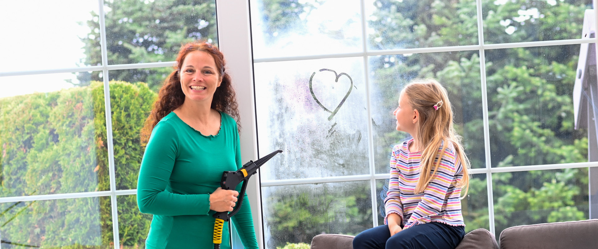 Ein kleines Mädchen malt mit dem Finger ein Herz auf die Fensterscheibe, die ihre Mutter gerade mit Trockendampf reinigt.